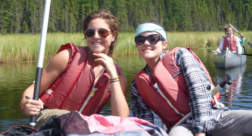 Two students sit in a watercraft and smile at the camera. Behind them is another canoe and the green shore.